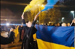 [ai] A group of people gathered at night, holding a large Ukrainian flag while some wave blue and yellow smoke flares. There are lights in the background and a crowd visible.