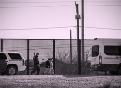 [ai] A Border Patrol officer walking alongside a child near a fenced border area, with two Border Patrol vehicles parked nearby and utility poles in the background.