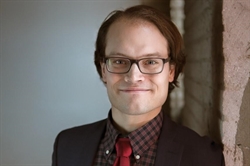 [ai] A man with glasses smiling at the camera, wearing a dark suit and a red tie over a checkered shirt, standing against a textured wall.