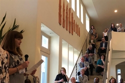 [ai] A speaker holding a microphone addresses an audience gathered on a staircase. People are seated and standing in a well-lit interior space decorated with artwork on the walls.