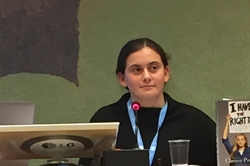 [ai] A young person, seated at a table, listening attentively during a presentation. A microphone is in front of them, with a glass of water nearby. Visible in the background is a book titled "I Have the Right To...".
