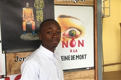 [ai] A young man in a white shirt standing against a poster displaying an eye with a teardrop and the text "NON A LA REINE DE MORT." Behind him, there is also a colorful poster of a figure in a creative costume.