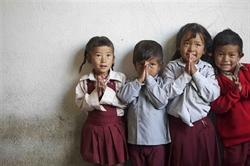 [ai] Four children standing in a line against a white wall, with their hands together in a prayer position. They are wearing school uniforms, consisting of shirts and skirts or shorts in various colors.