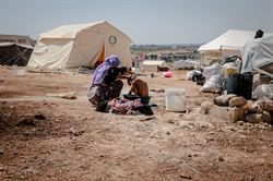[ai] A woman in a purple headscarf bathing a child in a basin outside in a makeshift camp with tents in the background. The ground is dry and dusty, with a few buckets and scattered belongings visible.