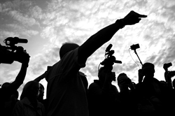 [ai] A silhouette of a person gesturing with one arm raised, surrounded by a crowd holding cameras. The sky is visible in the background with soft clouds.