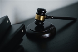 [ai] A wooden gavel with a gold band resting on a dark surface next to black binders. The background is softly blurred, focusing on the gavel.