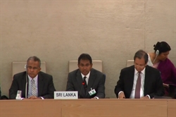 [ai] Three men seated at a conference table, with one man speaking into a microphone. A placard reading 'SRI LANKA' is visible in front of him. In the background, a woman wearing headphones is seated.