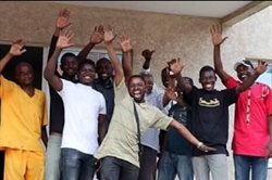 [ai] A group of eleven men standing together, smiling and raising their hands, in front of a building. They are wearing a mix of casual clothing in various colors.