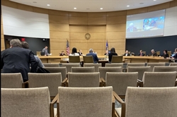 [ai] A view of a legislative meeting room with empty chairs in the foreground and several individuals seated around a circular table. A screen displays presentations, and flags are visible in the background.