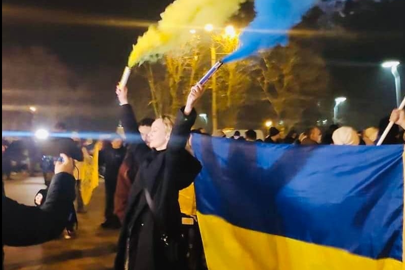 [ai] A group of people gathered at night, holding a large Ukrainian flag while some wave blue and yellow smoke flares. There are lights in the background and a crowd visible.