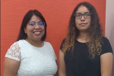 [ai] Two women standing side by side in front of a textured orange wall. The woman on the left is wearing a white lace top and glasses, while the woman on the right is in a black shirt with long curly hair.