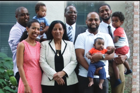 [ai] A group of six adults and three children posing together outdoors, with a house in the background. The adults are dressed in casual and formal attire. The children are being held or standing next to their guardians.