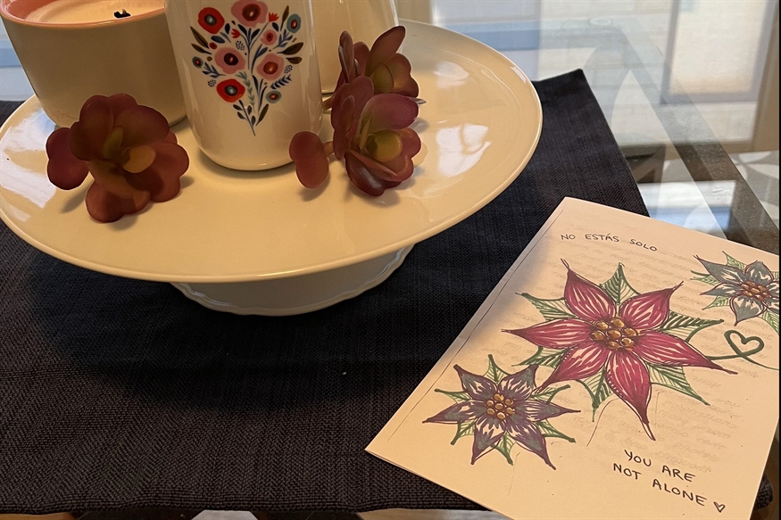 [ai] A decorative display featuring a white ceramic cup with floral designs, alongside small artificial flowers on a round white plate. A colorful card with hand-drawn flowers reads, 'YOU ARE NOT ALONE.'