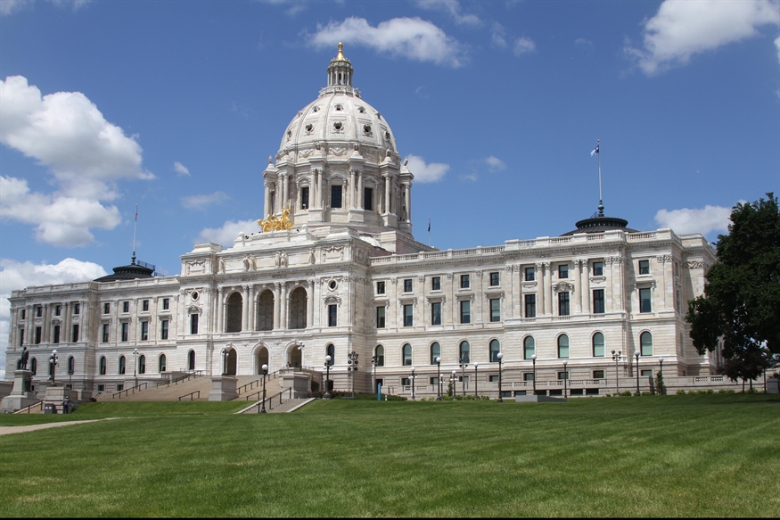 Exterior view of the Minnesota State Capitol building in St. Paul on a sunny day, featuring its white marble facade, grand staircase, central dome with a gold-leaf sculpture, and the new Minnesota state flag flying at the top