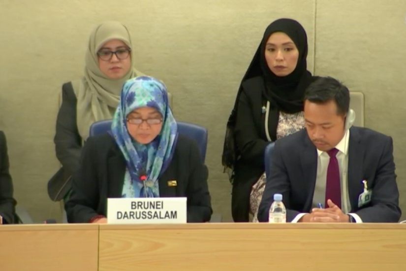 [ai] A woman wearing a hijab and glasses is speaking at a table labeled 'Brunei Darussalam' during a meeting, while two other women in hijabs and a man in a suit sit beside her in a formal setting.