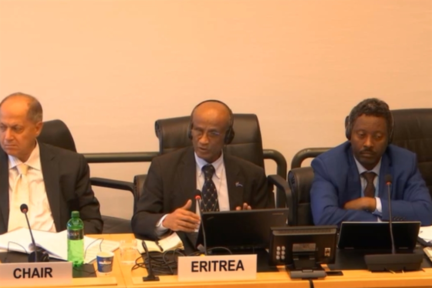 [ai] A panel discussion featuring three men seated at a table. The person in the center, labeled 'ERITREA', is speaking into a microphone while wearing headphones. Two additional panel members are seated on either side. A green beverage bottle is visible on the table.