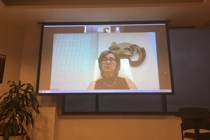 [ai] A woman with glasses, wearing a black top, appears on a large screen during a video call. She is seated in front of a light-colored wall with a decorative piece. A potted plant is visible in the foreground.