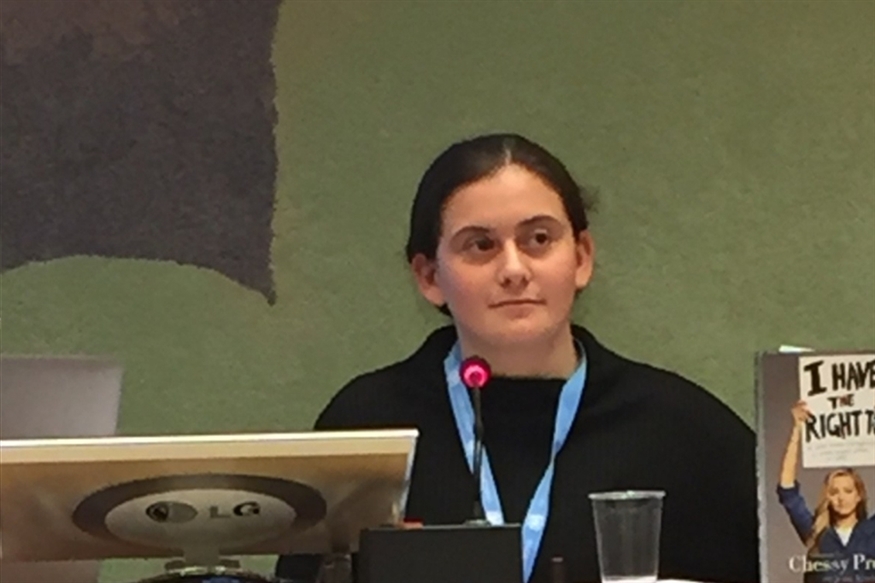 [ai] A young person, seated at a table, listening attentively during a presentation. A microphone is in front of them, with a glass of water nearby. Visible in the background is a book titled "I Have the Right To...".