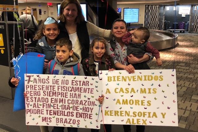 [ai] A group of five people, including a woman holding a child and four children, stands at an airport's baggage claim area. They are holding signs welcoming family members, with one sign in Spanish expressing love and reunion after several years.