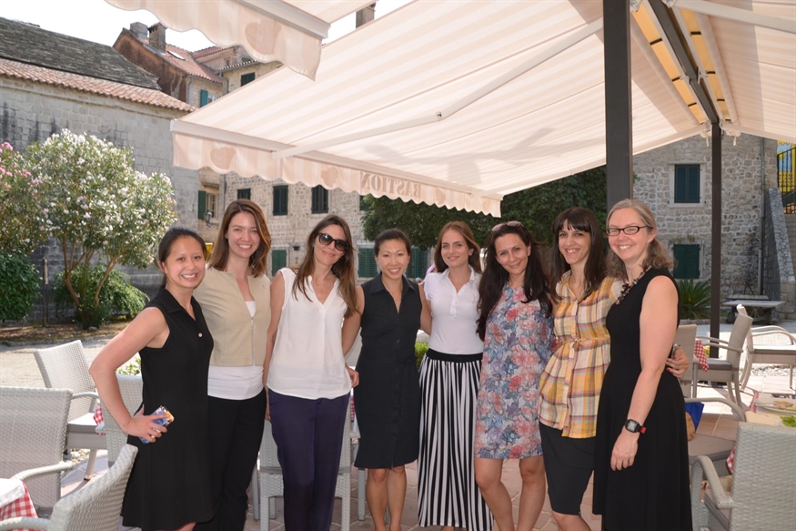 [ai] A group of eight women posing together outdoors under a canopy. They are smiling and dressed in various summer outfits, standing in an appealing outdoor setting with buildings and greenery in the background.
