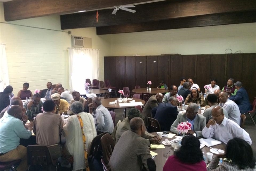 [ai] A large group of people seated at round tables in a well-lit room, engaged in discussion. Some individuals are taking notes while others are conversing. Centerpieces with flowers adorn the tables.