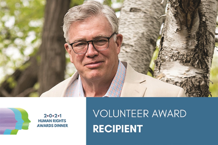 [ai] A man with glasses wearing a light-colored blazer stands outdoors among trees. On the left, there is a logo for a Human Rights Awards Dinner with the text '2021 Volunteer Award Recipient'.